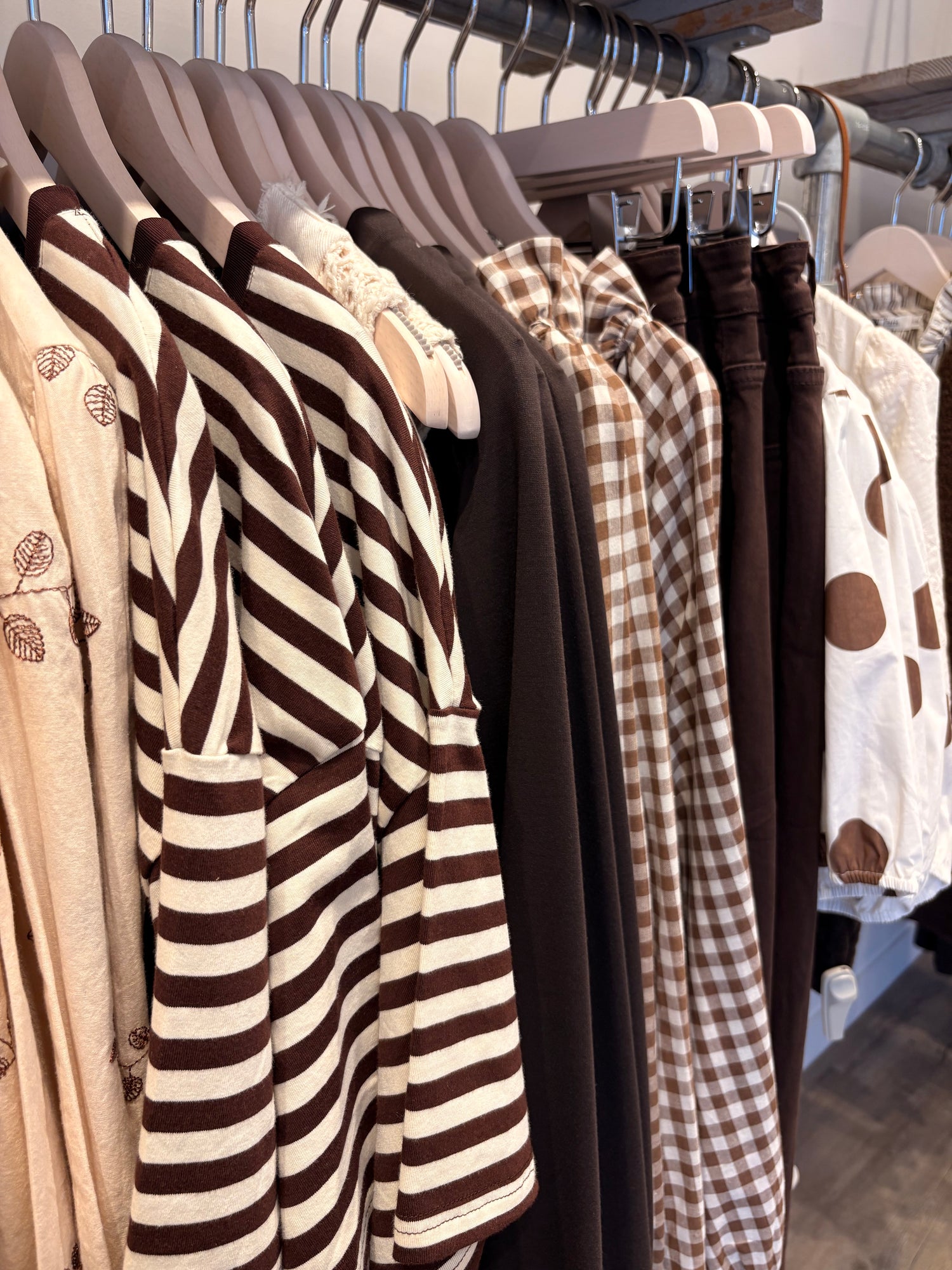 A close-up of a clothing rack displaying a variety of women's tops in earthy tones, including a prominent brown and cream striped shirt, a brown gingham top, and solid brown and white garments.